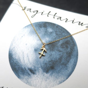 Peut inclure: Un collier en or avec un pendentif repr&eacute;sentant le signe du zodiaque Sagittaire. Le collier est sur une carte blanche avec le mot "Sagittaire" &eacute;crit en encre noire.