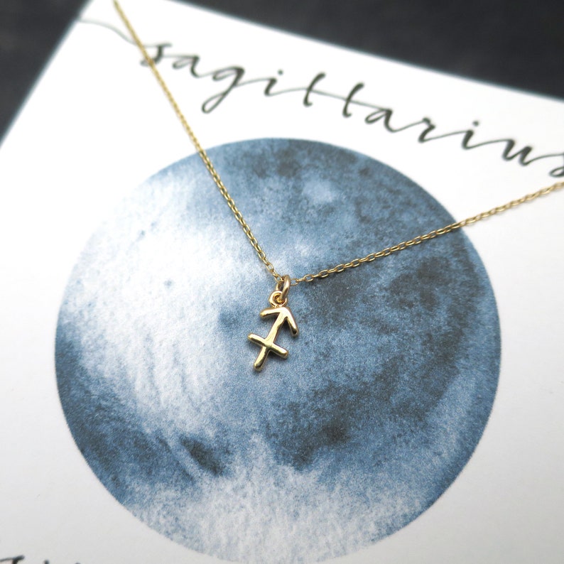 Peut inclure: Un collier en or avec un pendentif repr&eacute;sentant le signe du zodiaque Sagittaire. Le collier est sur une carte blanche avec le mot "Sagittaire" &eacute;crit en encre noire.