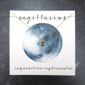 Peut inclure: Un collier avec un pendentif en or repr&eacute;sentant le signe du zodiaque Sagittaire, sur une carte blanche avec une lune en aquarelle bleue et le texte "sagittaire curieux &bull; optimiste".