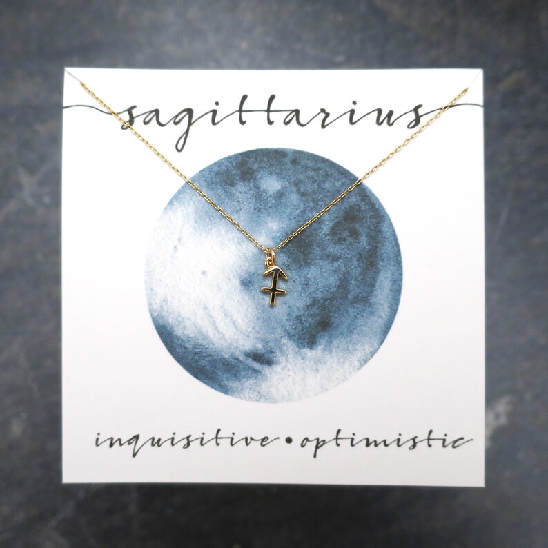 Peut inclure: Un collier avec un pendentif en or repr&eacute;sentant le signe du zodiaque Sagittaire, sur une carte blanche avec une lune en aquarelle bleue et le texte "sagittaire curieux &bull; optimiste".