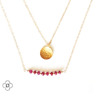 May include: A gold chain necklace with a hammered gold circle pendant and a second chain with a row of small red gemstones.