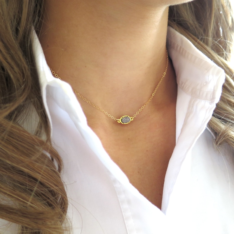 May include: A delicate gold chain necklace with a small, oval-shaped pendant featuring a raw, gray diamond.