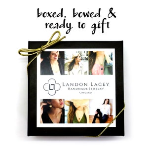 Peut inclure: Une bo&icirc;te cadeau noire avec un n&oelig;ud en ruban dor&eacute;. La bo&icirc;te a une &eacute;tiquette blanche avec le texte "LANDON LACEY HANDMADE JEWELRY CHICAGO" et un petit logo. La bo&icirc;te est entour&eacute;e d'images de femmes portant des colliers.