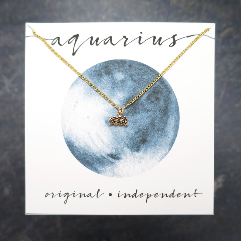 May include: A gold necklace with a small Aquarius zodiac sign charm on a white card with blue watercolor background. The card says "aquarius" and "original. independent".