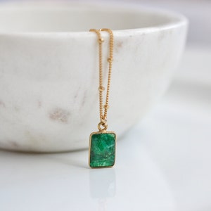Puede incluir: Un collar de cadena dorada con un colgante de piedra preciosa verde. El colgante tiene forma rectangular y una superficie texturizada.