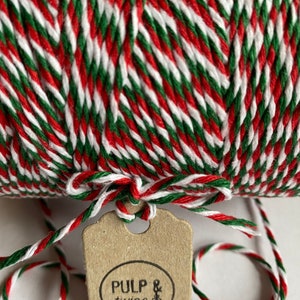 May include: A close-up of a spool of red, green, and white twine, with a kraft paper tag attached. The tag is tied with a knot and displays the words "Pulp & twine". The twine is tightly wound, creating a festive and decorative appearance.