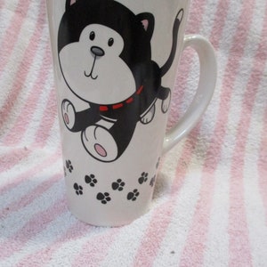 May include: White ceramic mug with a black and white cartoon cat design. The cat is wearing a red collar and has paw prints around it.