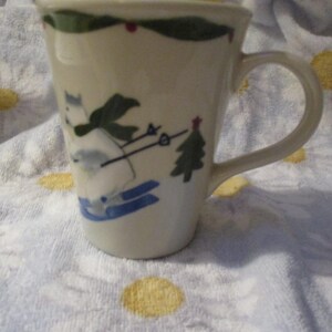 May include: A white ceramic mug featuring a green, blue and red winter scene. The scene shows a person skiing down a hill with a pine tree in the background.