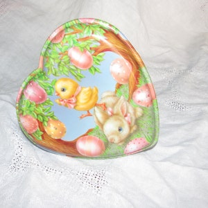 May include: A heart-shaped metal tin with a pastel blue background featuring a yellow chick and a white bunny rabbit. The tin is decorated with green leaves and pink and white eggs.