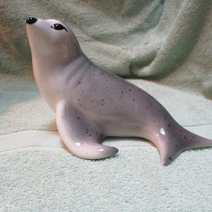 May include: A ceramic seal figurine with a grey and white speckled glaze. The seal is looking up with its mouth slightly open.