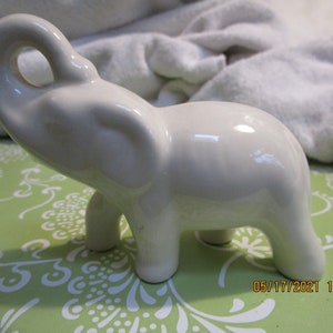 May include: A white ceramic elephant figurine with its trunk raised in the air. The elephant is standing on a green and white patterned surface.