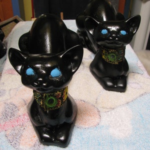 May include: Two black ceramic cat figurines with blue eyes and colourful patterned collars. The cats are sitting on a white fluffy surface.