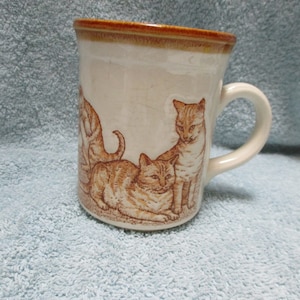 May include: A white ceramic mug with a brown rim and handle. The mug features a design of four orange tabby cats.