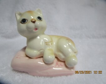 May include: A white ceramic figurine of a cat with brown markings, lying on a pink pillow. The cat has blue eyes and an open mouth.