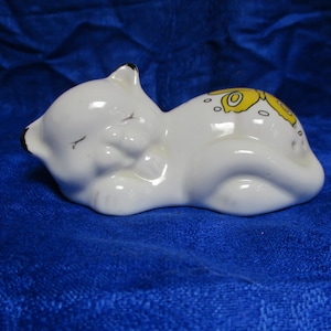 May include: A white ceramic cat figurine in a sleeping pose. The cat has closed eyes, black accents on the ears, and yellow floral designs on its back. The figurine is resting on a blue surface.