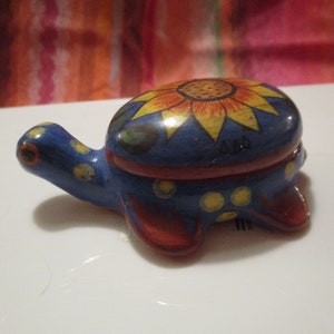 May include: A small blue ceramic turtle trinket box with a red and yellow sunflower design on the top. The turtle has yellow polka dots on its shell and red and yellow accents on its legs.