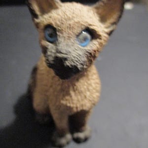 May include: A small figurine of a Siamese cat with blue eyes and a tan body. The cat is sitting upright, with its ears perked up. The figurine is detailed and appears to be made of a hard material.