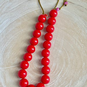 May include: A vibrant red beaded necklace with large, round beads. The necklace is strung on a gold-colored cord and features a small, pink bead near the clasp. The beads are evenly spaced and have a glossy finish.