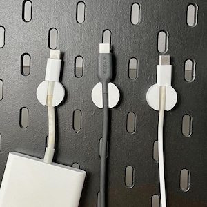 May include: Three white cable organizers with black cables attached to them are mounted on a black pegboard. The cable organizers have a white circle in the center. The cables are hanging down from the organizers. One of the cables is connected to a white adapter.