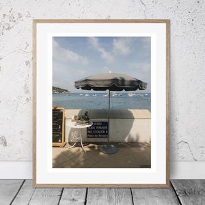 May include: Framed photograph of a beach scene with a dark gray umbrella, small white table, and a sign with French text. The ocean and boats are visible in the background under a blue sky. A small chalkboard is on the left.