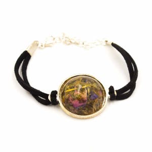 May include: A bracelet featuring a clear, round centerpiece filled with dried flowers in shades of pink, purple, and green. The centerpiece is encased in a silver-colored metal setting. The bracelet has a black suede cord and a silver-colored chain.
