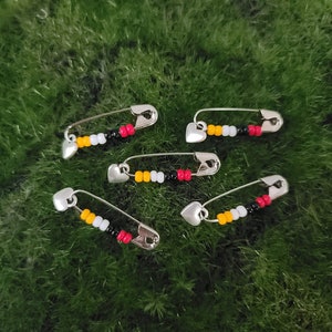 May include: Five safety pins with heart-shaped tops decorated with colorful beads. The beads are black, white, yellow, and red.
