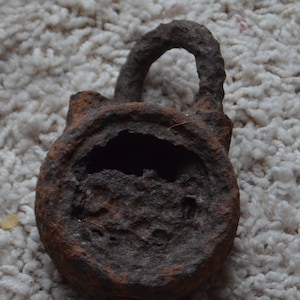 Puede incluir: Un candado de metal oxidado con un diseño circular y una cerradura. Al candado le falta una parte del metal en la parte delantera.