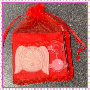 Op de afbeelding: Een rode organza geschenktas met een lichtroze gezichtsvormige zeep, twee kleine verfpotjes met rode en roze verf en een dunne penseel. De tas is vastgebonden met een rood lint.