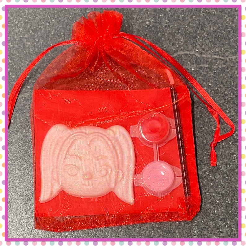 Op de afbeelding: Een rode organza geschenktas met een lichtroze gezichtsvormige zeep, twee kleine verfpotjes met rode en roze verf en een dunne penseel. De tas is vastgebonden met een rood lint.