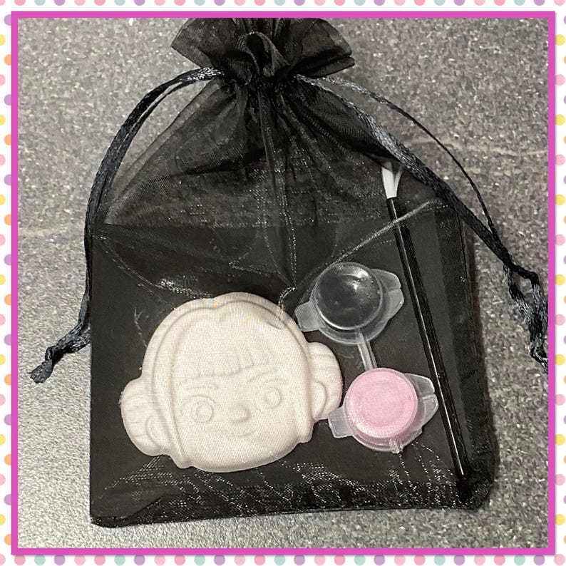 Op de afbeelding: Een zwarte organza geschenktas met een wit, cartoonachtig gezichtsvormig item, een doorzichtige plastic schep, een roze plastic bakje en een zwarte stok. De tas is vastgebonden met een zwart lint.