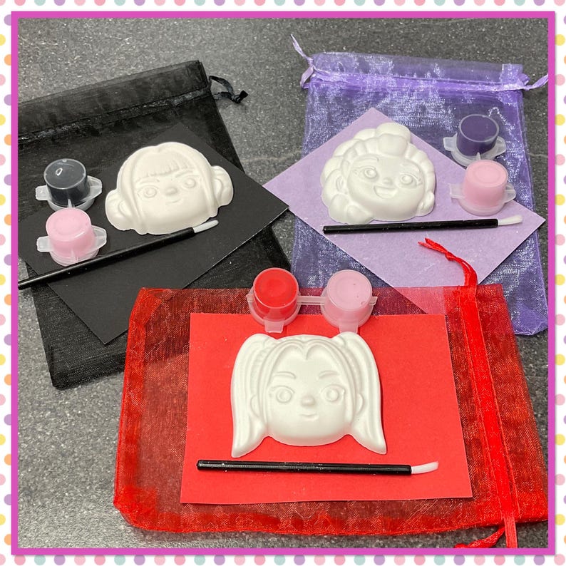 Op de afbeelding: Drie doe-het-zelf schilder kits met witte gipsen gezichtsvormen, verfpotten en penselen. Elke kit is verpakt in een andere gekleurde zak: zwart, paars en rood. De gezichtsvormen hebben verschillende kapsels.