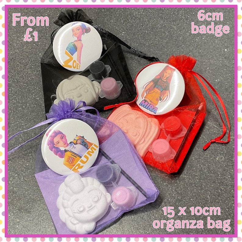 Op de afbeelding: Drie organza zakjes, elk met een badge van 6 cm, een kleine beschilderbare figuur en verfpotjes. De badges tonen illustraties van personages genaamd Zoe, Mira en Rumi. De zakjes zijn zwart, rood en paars.