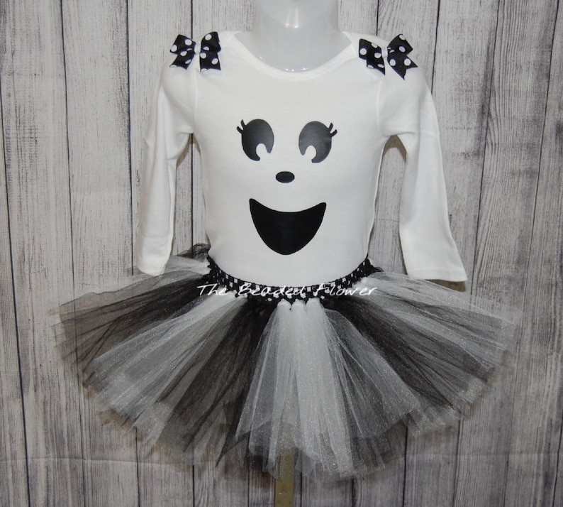 May include: A white long-sleeved shirt with a black ghost face design and black polka dot bows on the shoulders. The shirt is paired with a black and white tulle tutu skirt.