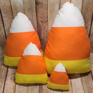 May include: Four plush pillows shaped like candy corn. The pillows are in various sizes and are made of white, orange, and yellow fabric.