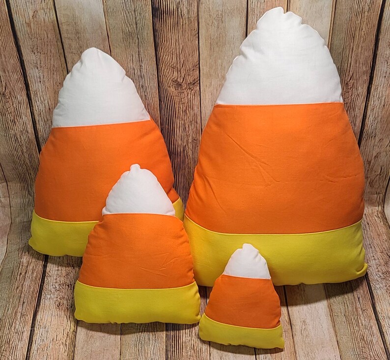 May include: Four plush pillows shaped like candy corn. The pillows are in various sizes and are made of white, orange, and yellow fabric.