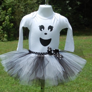 May include: A white long-sleeved onesie with a black ghost face design. The onesie is paired with a black and white tulle tutu with a black polka dot ribbon.