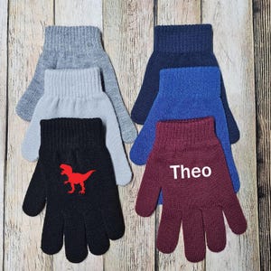 May include: A collection of five knit gloves in gray, light gray, black, navy blue, and maroon. The black glove features a red dinosaur graphic, while the maroon glove has the white text "Theo" printed on the back of the hand area.