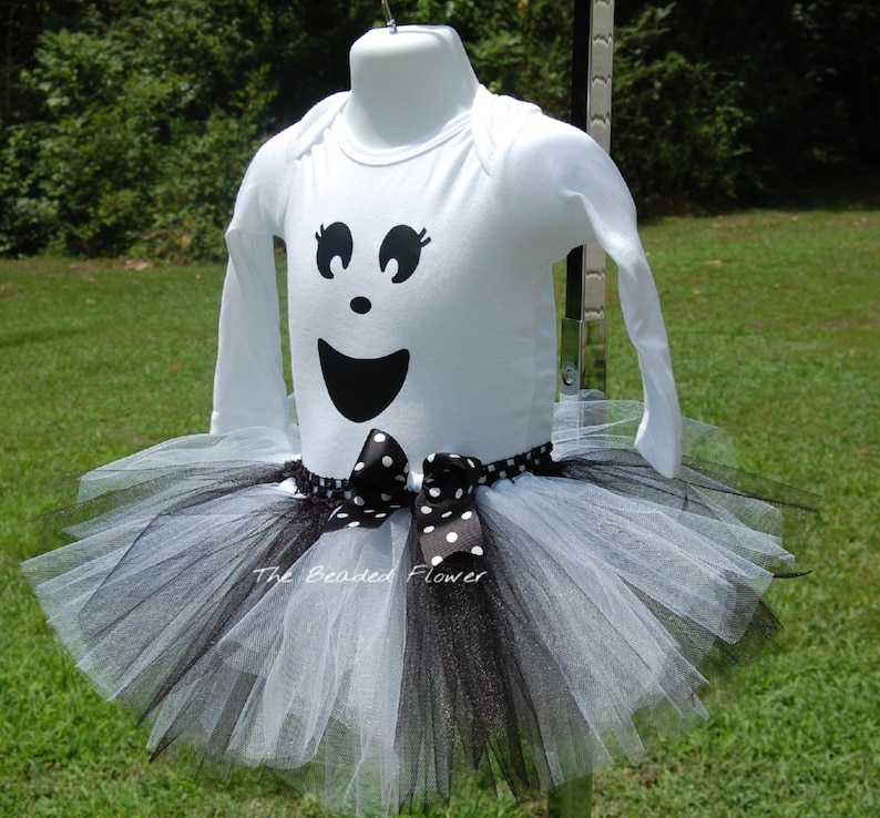 May include: White long-sleeved shirt with a black ghost face design and a black and white tulle tutu with a black polka dot bow.