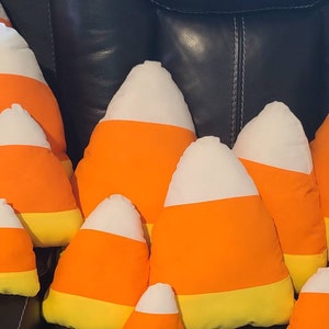 May include: A group of plush candy corn pillows in various sizes, arranged on a black leather couch. The pillows are made of soft fabric and feature a white, orange, and yellow color scheme.