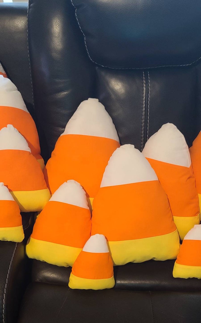 May include: A group of plush candy corn pillows in various sizes, arranged on a black leather couch. The pillows are made of soft fabric and feature a white, orange, and yellow color scheme.