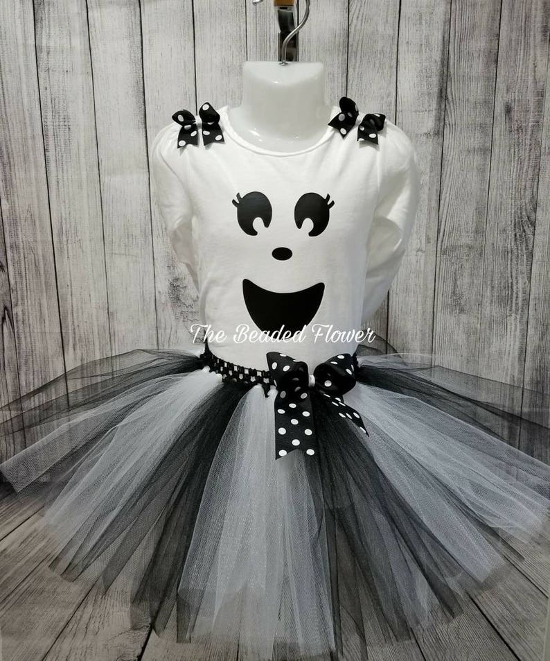 May include: White long-sleeved shirt with a black ghost face design and black polka dot bows on the shoulders. A black and white tulle tutu with a black polka dot bow.