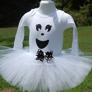 May include: White tutu with a white long-sleeved shirt featuring a black ghost face with a black and white polka dot bow. The text "The Beaded Flower" is visible on the bottom of the shirt.
