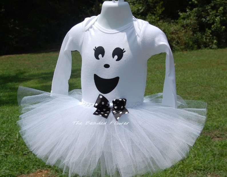 May include: White tutu with a white long-sleeved shirt featuring a black ghost face with a black and white polka dot bow. The text "The Beaded Flower" is visible on the bottom of the shirt.