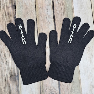 May include: A pair of black knit gloves with the word "BITCH" printed in white on the back of each hand. The gloves are displayed on a light brown wooden surface.