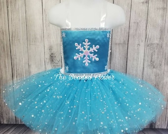 May include: A blue tulle tutu with silver glitter and a blue bodice with a white snowflake design and rhinestones. The bodice has white straps.