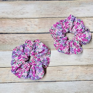 May include: Two purple and pink floral print scrunchies. The scrunchies are made of fabric and have a gathered look.