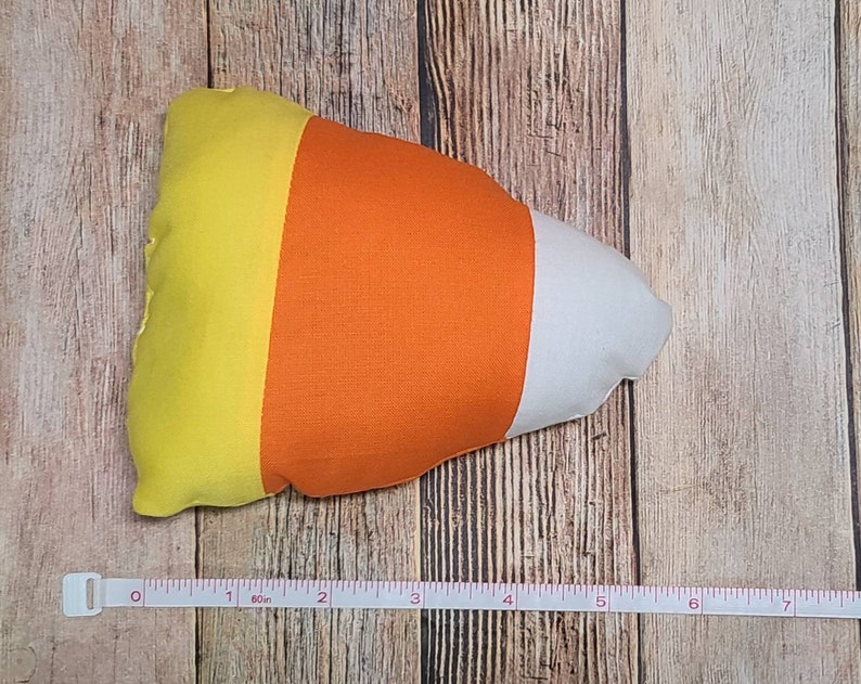 May include: A yellow, orange, and white candy corn shaped pillow. The pillow is about 7 inches long.