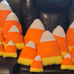 May include: A group of 10 plush candy corn pillows in various sizes, arranged in a pyramid shape on a brown leather couch. The pillows are made of soft fabric and feature the classic candy corn colors of orange, yellow, and white.