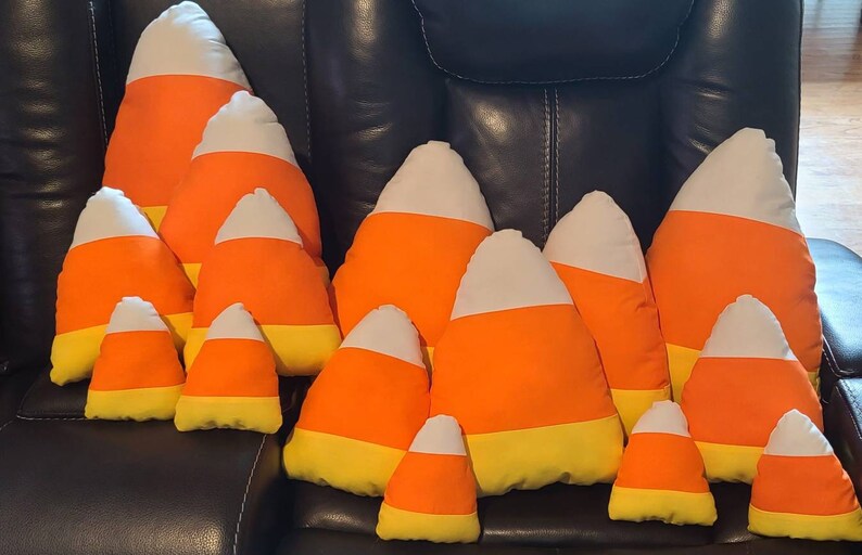 May include: A group of 10 plush candy corn pillows in various sizes, arranged in a pyramid shape on a brown leather couch. The pillows are made of soft fabric and feature the classic candy corn colors of orange, yellow, and white.