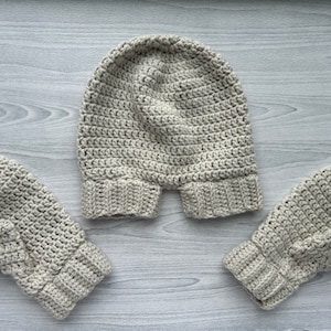 May include: A set of crocheted winter accessories in a light beige colour. The set includes a hat and two mittens.